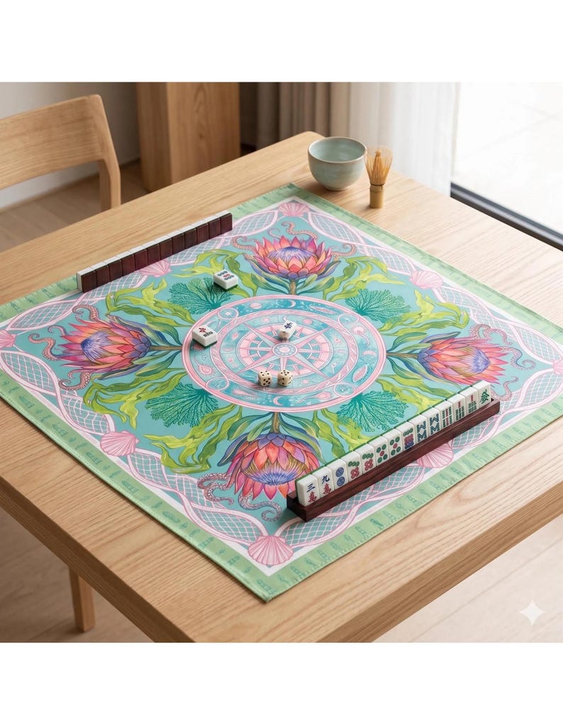 May include: A square Mahjong game mat with a floral design in shades of teal, pink, and green. The mat features a central compass rose design and is surrounded by decorative borders. Brown tile racks and game tiles are arranged on the mat.
