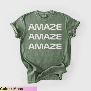 May include: A moss-colored t-shirt with the word "AMAZE" printed three times in white, stacked vertically. The shirt has a crew neck and short sleeves. The fabric appears soft and slightly faded.