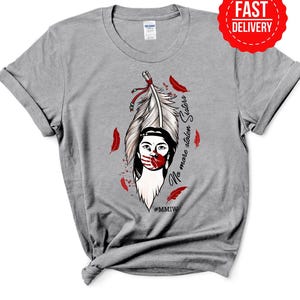 May include: A gray t-shirt featuring a graphic of a Native American woman with a feather headdress and red handprint. The text "No more stolen Sisters" and the hashtag #MMIW are also visible. A red "Fast Delivery" sticker is on the top right.