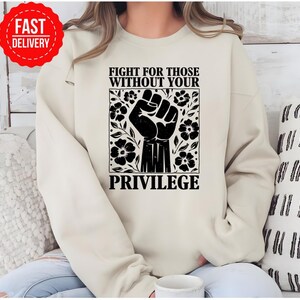 May include: Cream-colored sweatshirt with a black graphic of a raised fist surrounded by flowers and the text "FIGHT FOR THOSE WITHOUT YOUR PRIVILEGE." A red "FAST DELIVERY" sticker is in the upper left corner. The sweatshirt is a casual garment.