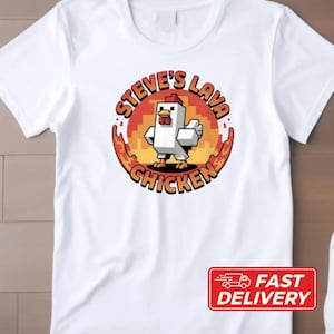 May include: White t-shirt featuring a cartoon chicken character with the text "Steve's Lava Chicken" in a circular design. The design includes red, orange, and yellow colors. A red "Fast Delivery" sticker is in the bottom right corner.