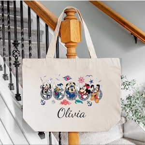May include: A white canvas tote bag with a nautical theme featuring Mickey Mouse and friends. The bag has a black handle and the name "Olivia" printed on the front.