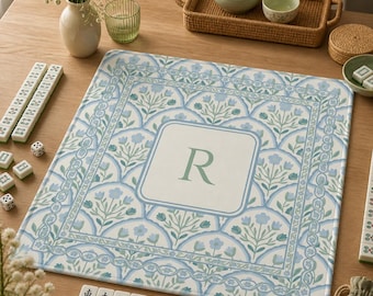 May include: A square, floral-patterned mahjong mat in shades of blue and white, with a central square featuring the letter "R". The mat is surrounded by mahjong tiles, dice, and a small vase of flowers. A tea set is in the background.
