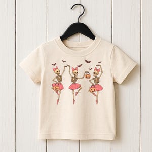 May include: Cream-colored t-shirt featuring a Halloween-themed graphic. The design shows three skeleton ballerinas wearing pink tutus and bows, holding pumpkin-shaped treat bags, with bats flying above. The shirt is hanging on a black hanger.