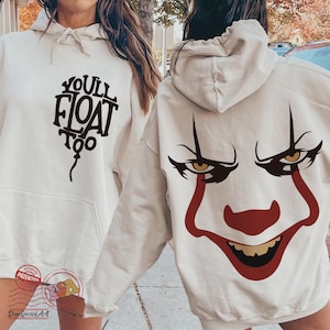 May include: Beige hoodie with a front pocket and drawstring hood. The front features the text "YOU'LL FLOAT TOO" in a black, stylized font. The back displays a red and black graphic of a clown's face with yellow eyes.