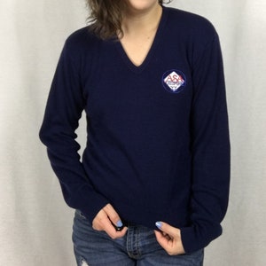 May include: A navy blue long-sleeved sweater with a v-neck and a small embroidered patch on the left chest. The patch is red, white, and blue and says "ASA Olympics".