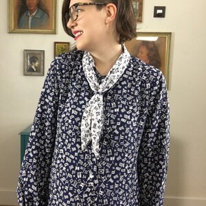 May include: A navy blue blouse with a white and black pattern of Roman numerals and flags. The blouse has a white scarf tied around the neck.