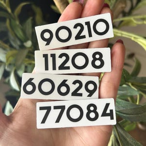 May include: Four white rectangular tags with black numbers: 90210, 11208, 60629, and 77084. The tags are held in a hand, with a background of green leaves and a brown stem.