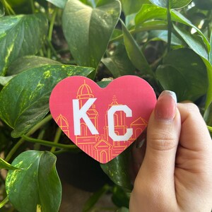 K&ouml;nnte beinhalten: Ein roter herzf&ouml;rmiger Aufkleber mit einem wei&szlig;en "KC" in der Mitte. Der Aufkleber hat einen gelben Umriss und ein Design von Geb&auml;uden im Herzen.