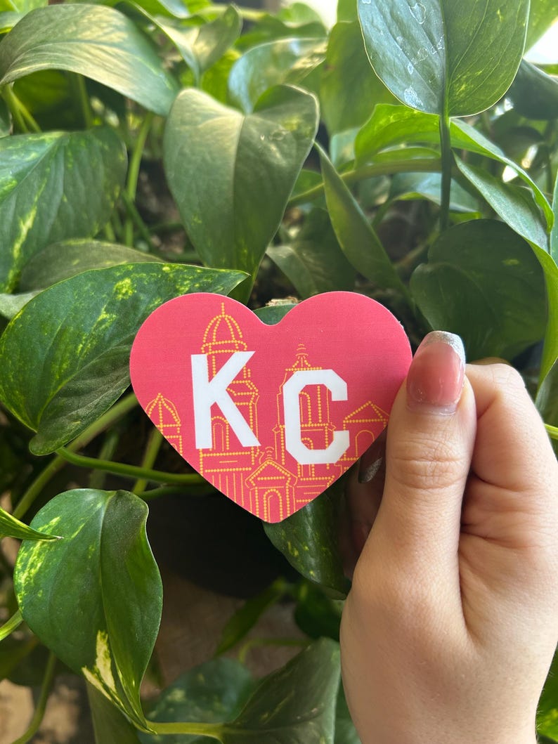 K&ouml;nnte beinhalten: Ein roter herzf&ouml;rmiger Aufkleber mit einem wei&szlig;en "KC" in der Mitte. Der Aufkleber hat einen gelben Umriss und ein Design von Geb&auml;uden im Herzen.