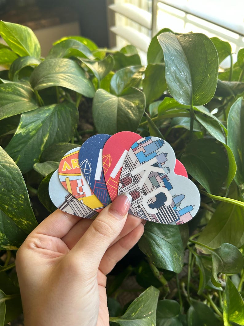 May include: Five heart-shaped stickers with different cityscapes printed on them. The stickers are in a hand, and the background is a green leafy plant.