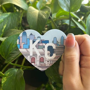 K&ouml;nnte beinhalten: Ein herzf&ouml;rmiger Aufkleber mit einer bunten Illustration der Skyline von Kansas City, einschlie&szlig;lich des Union Station-Geb&auml;udes. Der Aufkleber tr&auml;gt den Text "KC" in Wei&szlig;.