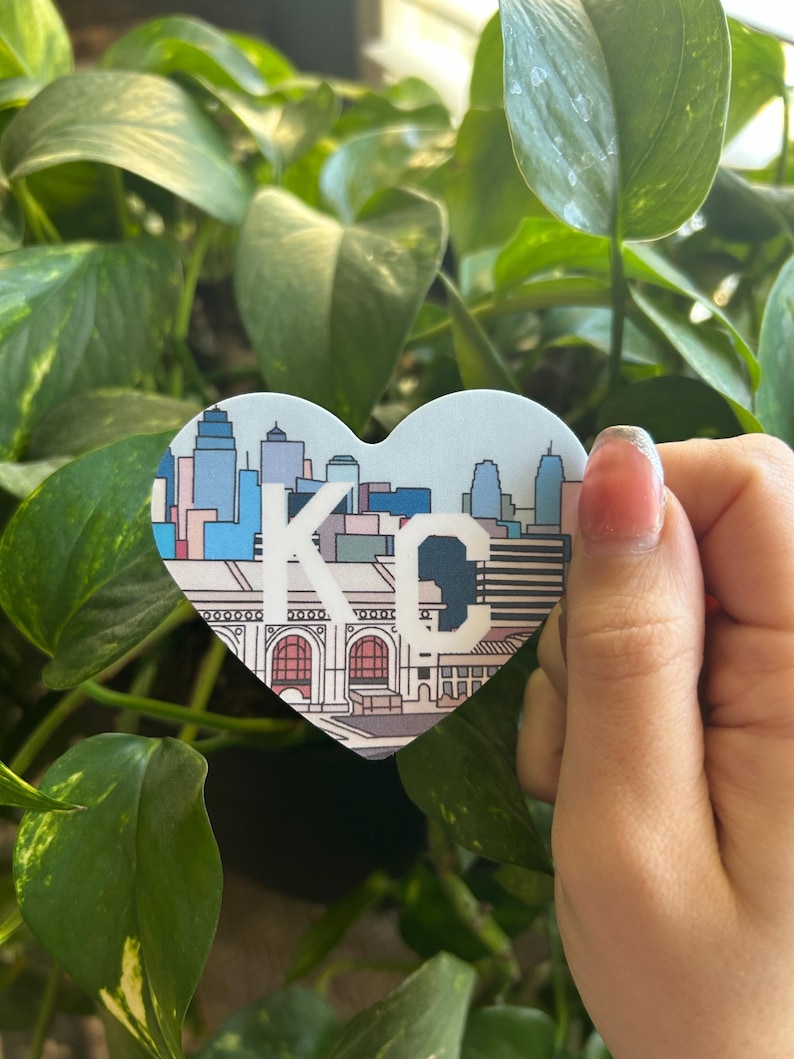 K&ouml;nnte beinhalten: Ein herzf&ouml;rmiger Aufkleber mit einer bunten Illustration der Skyline von Kansas City, einschlie&szlig;lich des Union Station-Geb&auml;udes. Der Aufkleber tr&auml;gt den Text "KC" in Wei&szlig;.