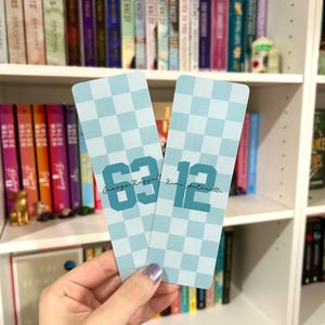 May include: Two light blue checkered pattern bookmarks with the numbers "63" and "12" in teal. The names "George Russell" and "Kimi Antonelli" are printed above the numbers. The background shows a bookshelf filled with books.