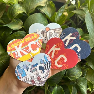K&ouml;nnte beinhalten: F&uuml;nf herzf&ouml;rmige Aufkleber mit verschiedenen Designs, die die Buchstaben "KC" und verschiedene Sehensw&uuml;rdigkeiten von Kansas City zeigen. Die Aufkleber werden in einer Hand gehalten.