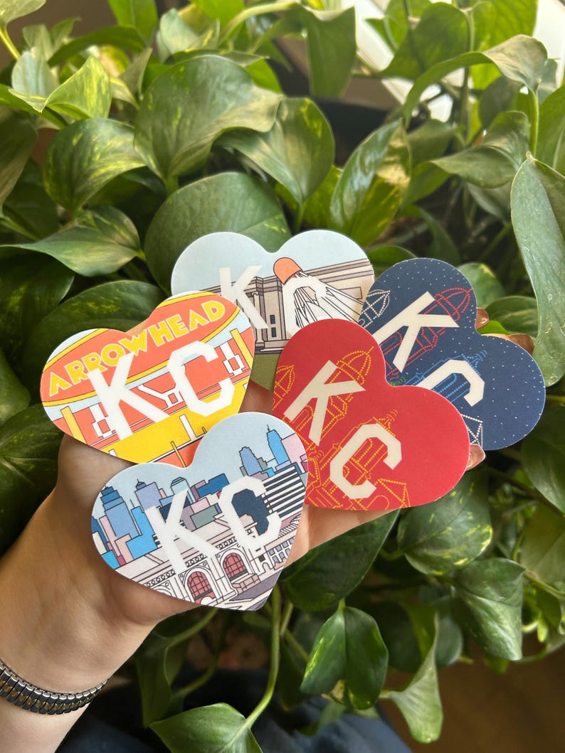 K&ouml;nnte beinhalten: F&uuml;nf herzf&ouml;rmige Aufkleber mit verschiedenen Designs, die die Buchstaben "KC" und verschiedene Sehensw&uuml;rdigkeiten von Kansas City zeigen. Die Aufkleber werden in einer Hand gehalten.