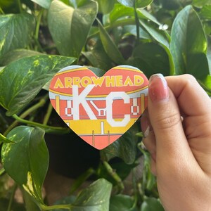 K&ouml;nnte beinhalten: Ein herzf&ouml;rmiger Aufkleber mit den Worten "Arrowhead KC" in wei&szlig;en Buchstaben auf gelbem Hintergrund. Der Aufkleber ist eine stilisierte Darstellung eines Fu&szlig;ballstadions.