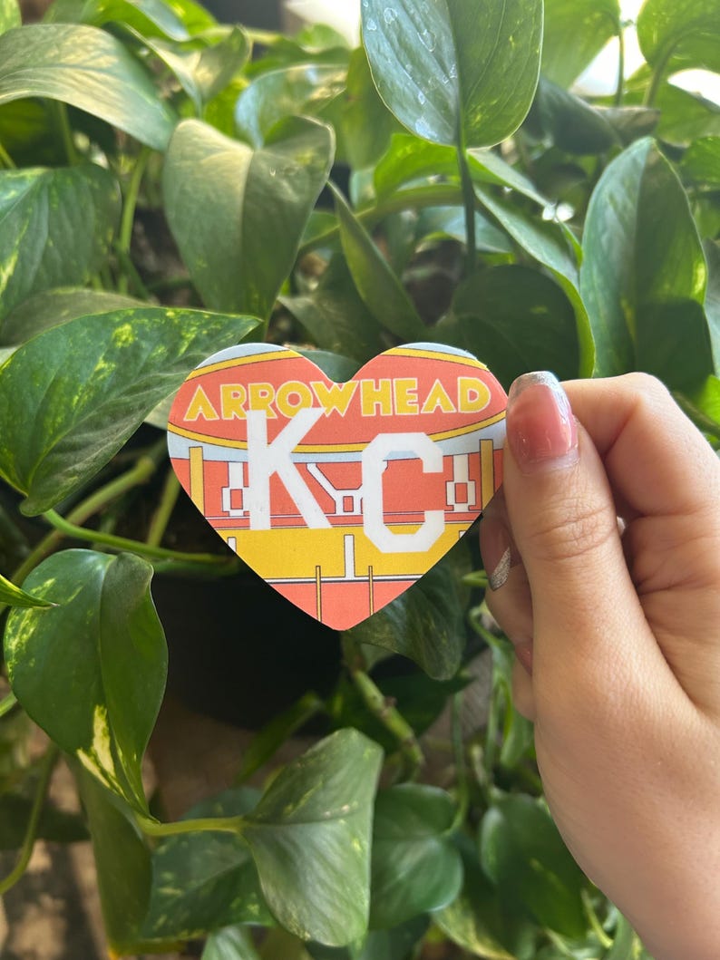 K&ouml;nnte beinhalten: Ein herzf&ouml;rmiger Aufkleber mit den Worten "Arrowhead KC" in wei&szlig;en Buchstaben auf gelbem Hintergrund. Der Aufkleber ist eine stilisierte Darstellung eines Fu&szlig;ballstadions.