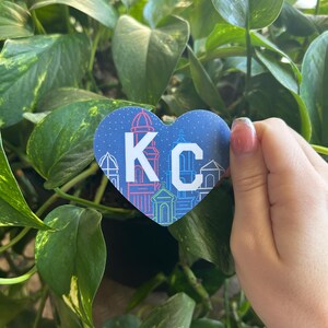 K&ouml;nnte beinhalten: Ein blauer herzf&ouml;rmiger Aufkleber mit wei&szlig;em Rand und den Buchstaben "KC" in Wei&szlig; mit blauem Rand. Der Aufkleber hat ein Stadtbilddesign mit Geb&auml;uden und Lichtern.