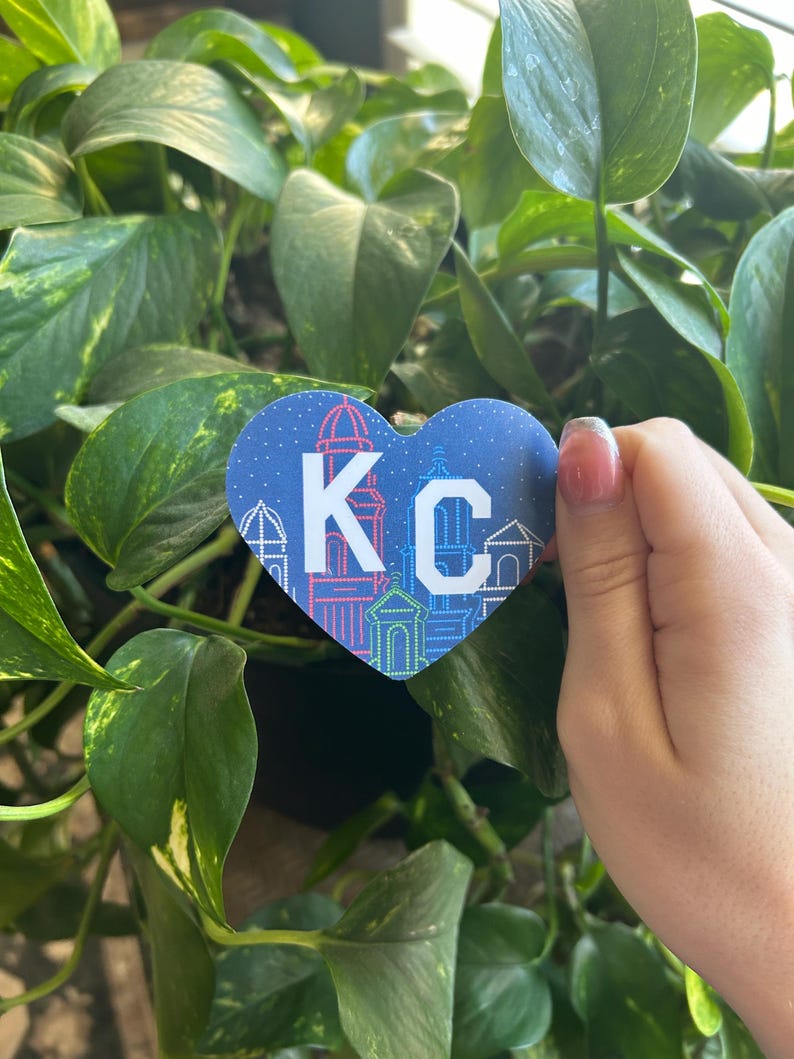 K&ouml;nnte beinhalten: Ein blauer herzf&ouml;rmiger Aufkleber mit wei&szlig;em Rand und den Buchstaben "KC" in Wei&szlig; mit blauem Rand. Der Aufkleber hat ein Stadtbilddesign mit Geb&auml;uden und Lichtern.