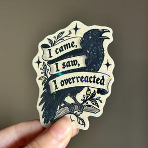 Pode incluir: Um adesivo recortado com um corvo preto pousado num galho, sobreposto por uma fita cor de creme exibindo o texto preto I came, I saw, I overreacted. O design inclui detalhes de estrelas holográficas por toda a ilustração.