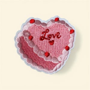 May include: Embroidered heart-shaped patch in shades of pink, with the word "Love" in red script. The patch features a layered cake design with red accents and a scalloped edge, perfect for adding to clothing or accessories.