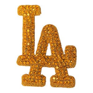 May include: A gold-colored, bedazzled "LA" logo, likely for the Los Angeles Dodgers. The letters are encrusted with small, round, golden rhinestones, creating a sparkling effect. The logo is set against a plain white background.