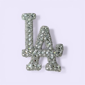 May include: A silver-colored, bedazzled "LA" logo, likely a team emblem. The letters are encrusted with small, reflective stones, creating a sparkling effect. The background is a soft, light purple.