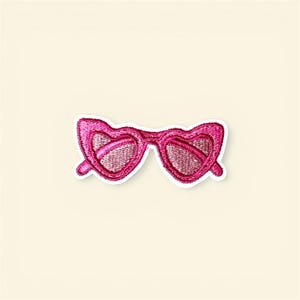 May include: A pink embroidered patch in the shape of heart-shaped sunglasses. The patch has a white border and a textured appearance. The lenses are filled with a mesh-like design.