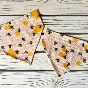 Puede incluir: Dos pañuelos triangulares con un estampado de abejas. La tela presenta un diseño de panal con abejas negras y formas de panal amarillas sobre un fondo rosa claro. Los pañuelos se muestran sobre una superficie de madera blanca.