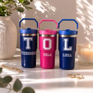 May include: Three insulated tumblers with carrying handles. Two are dark blue and one is bright pink. Each features a large white varsity-style letter and a name printed below: Theodore, Orla, and Luca. The tumblers have silver metal rims.