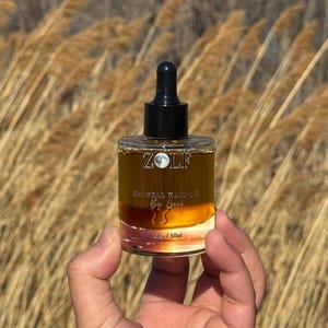 May include: A clear glass bottle with a black dropper top and a gold label that reads "ZOLF NATURAL HAIR OIL by Luna" and "1 fl oz / 50ml".