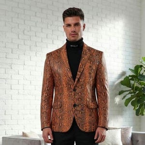 Puede incluir: Un blazer con estampado de piel de serpiente en color cobre y negro con dos botones frontales y solapas de muesca, usado sobre un suéter negro de cuello alto con el texto BAALLD en el cuello, combinado con pantalones negros y botines de cuero negro.