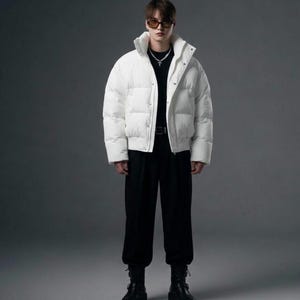 May include: A white puffer jacket with a high collar, front snap buttons, and long sleeves. The jacket is worn over a black shirt and black trousers, paired with black boots. A silver cross necklace and dark-tinted glasses complete the outfit.