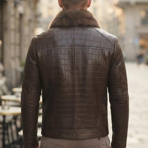 May include: A dark brown jacket with a textured crocodile skin pattern and a thick, plush fur collar. The jacket features long sleeves, a straight hemline, and visible stitching details across the back panel, worn over light brown trousers.