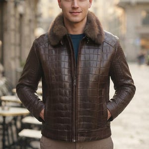 May include: A dark brown jacket with a textured crocodile-skin pattern and a thick, fluffy brown fur collar. The jacket features a central vertical zipper, long sleeves, and two side pockets. It is worn over a dark teal shirt and tan trousers.