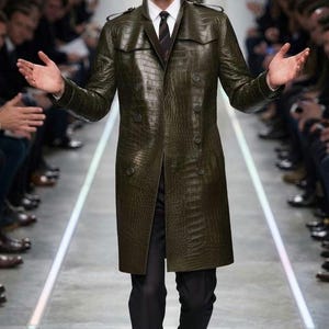 May include: A dark olive green trench coat with a textured crocodile skin pattern, featuring a double-breasted button closure, epaulets on the shoulders, and a high collar, worn over a white collared shirt, a dark striped tie, and black trousers.