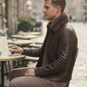 May include: A dark brown jacket with a textured crocodile-embossed pattern and a plush brown fur collar, paired with light brown trousers. The jacket features a front zipper closure and long sleeves, providing a structured look for the wearer.