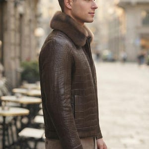 May include: A dark brown leather jacket with a textured crocodile-skin pattern and a thick, plush brown fur collar. The jacket features a front zipper closure, long sleeves with cuffs, and a straight hemline, worn with light brown trousers.