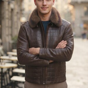 May include: A dark brown leather jacket with a textured crocodile-skin pattern, featuring a thick, plush brown fur collar and a central zipper closure. The jacket is worn over a dark blue shirt, paired with light brown trousers in a neutral tone.