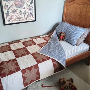 May include: A child's bedroom scene featuring a wooden bed with a quilt in red, brown, and white star patterns. A framed vintage bird chart hangs on the wall. A blue striped pillow and a toy are on the bed. A wooden pull-along toy is on the floor.