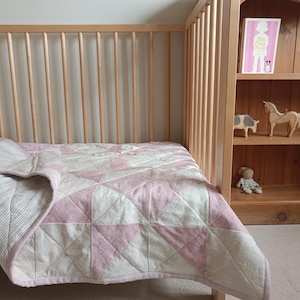 Puede incluir: Una cuna con un edredón con estampado geométrico rosa y blanco. El edredón está doblado sobre el lado de la cuna. En el fondo se ve una estantería de madera, con objetos decorativos en los estantes.