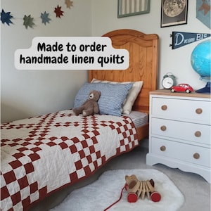 May include: A handmade linen quilt featuring a rust-coloured and off-white checkered pattern draped over a wooden bed frame. The quilt displays the text Made to order handmade linen quilts. The bed includes a blue and white striped pillow.