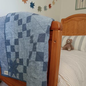 May include: A patchwork quilt featuring a blue and white colour palette with a mix of solid blue fabric squares and blue-and-white striped fabric squares. The quilt has a rectangular shape and is draped over the wooden footboard of a bed.