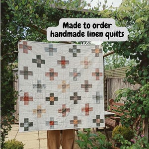 May include: A square linen quilt featuring a grid pattern of multicoloured plus signs in shades of terracotta, charcoal, and cream on an off-white background. The text "Made to order handmade linen quilts" is printed in black on a white speech bubble.