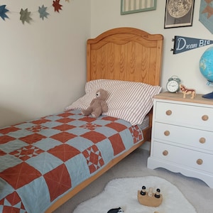 May include: A child's bedroom with a wooden bed, a quilt with a blue and rust-colored geometric pattern, and a white dresser. A teddy bear sits on the pillow. Decorative star-shaped garland hangs on the wall. A white rug with wooden toys.