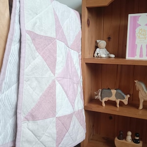 Puede incluir: Una colcha para bebé con un patrón geométrico rosa y blanco cuelga sobre una cuna. La colcha tiene un borde rosa y un reverso a rayas. Una estantería de madera exhibe juguetes y un libro.