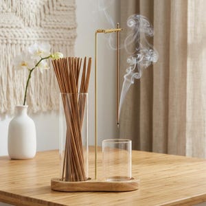 Peut inclure: Une base en bois supporte un grand cylindre en verre rempli de bâtonnets d'encens bruns et un cylindre en verre plus court et vide. Une fine tige en métal doré se dresse entre eux, avec une pince au sommet tenant un bâtonnet d'encens allumé.