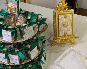 Puede incluir: Una exhibición escalonada de pequeñas cajas transparentes llenas de golosinas, atadas con cintas verdes y adornadas con detalles de perlas. También hay un libro decorativo con marco dorado y una tarjeta decorativa blanca y dorada.