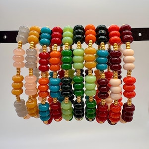 May include: A collection of beaded bracelets in various colors, including gray, yellow, pink, red, orange, teal, green, black, and burgundy. Each bracelet features round beads and gold-colored spacers, strung together. The bracelets are displayed on a black bar.